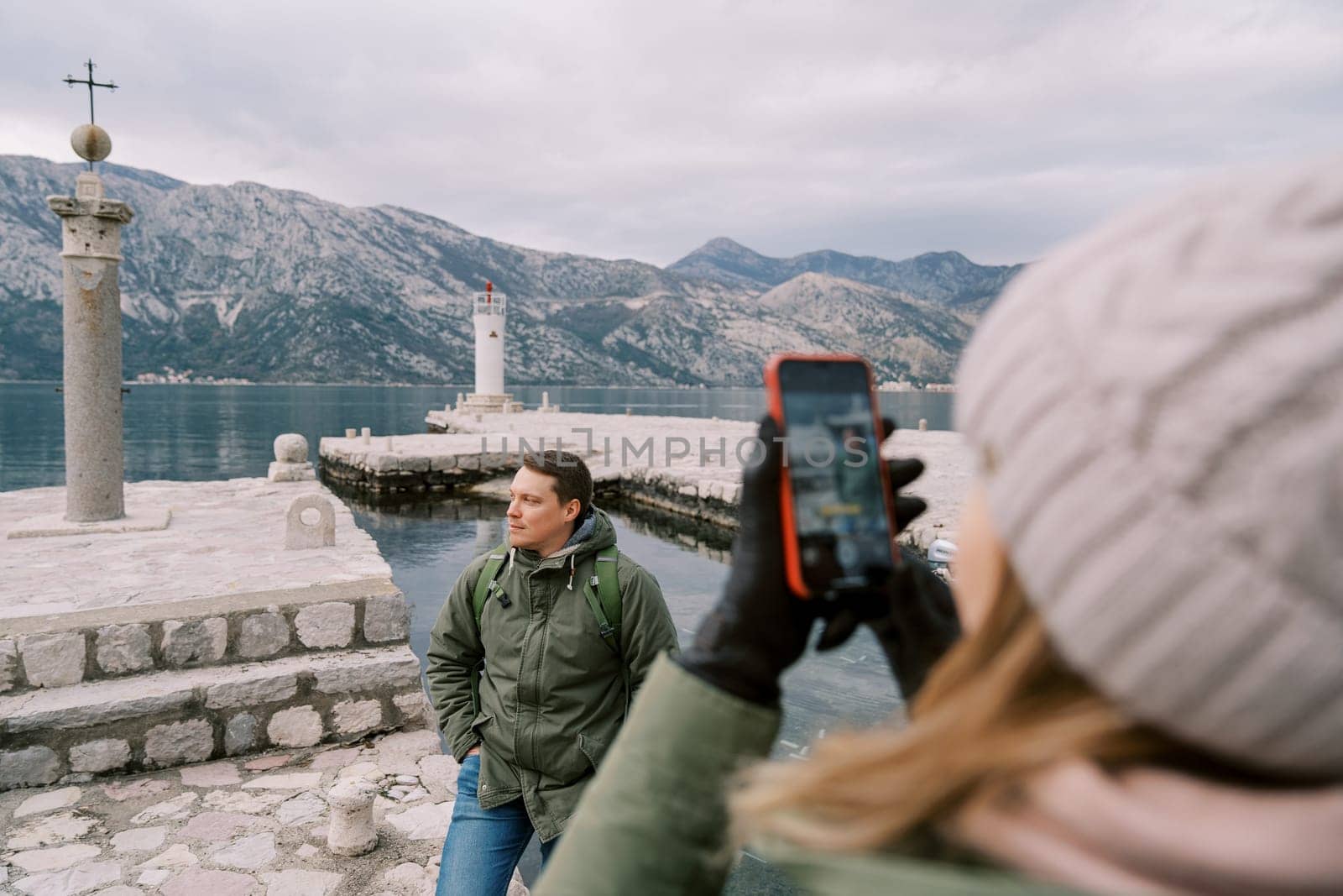 Woman takes a picture with a smartphone of a man standing on a pier near a column with a cross. Back view. High quality photo