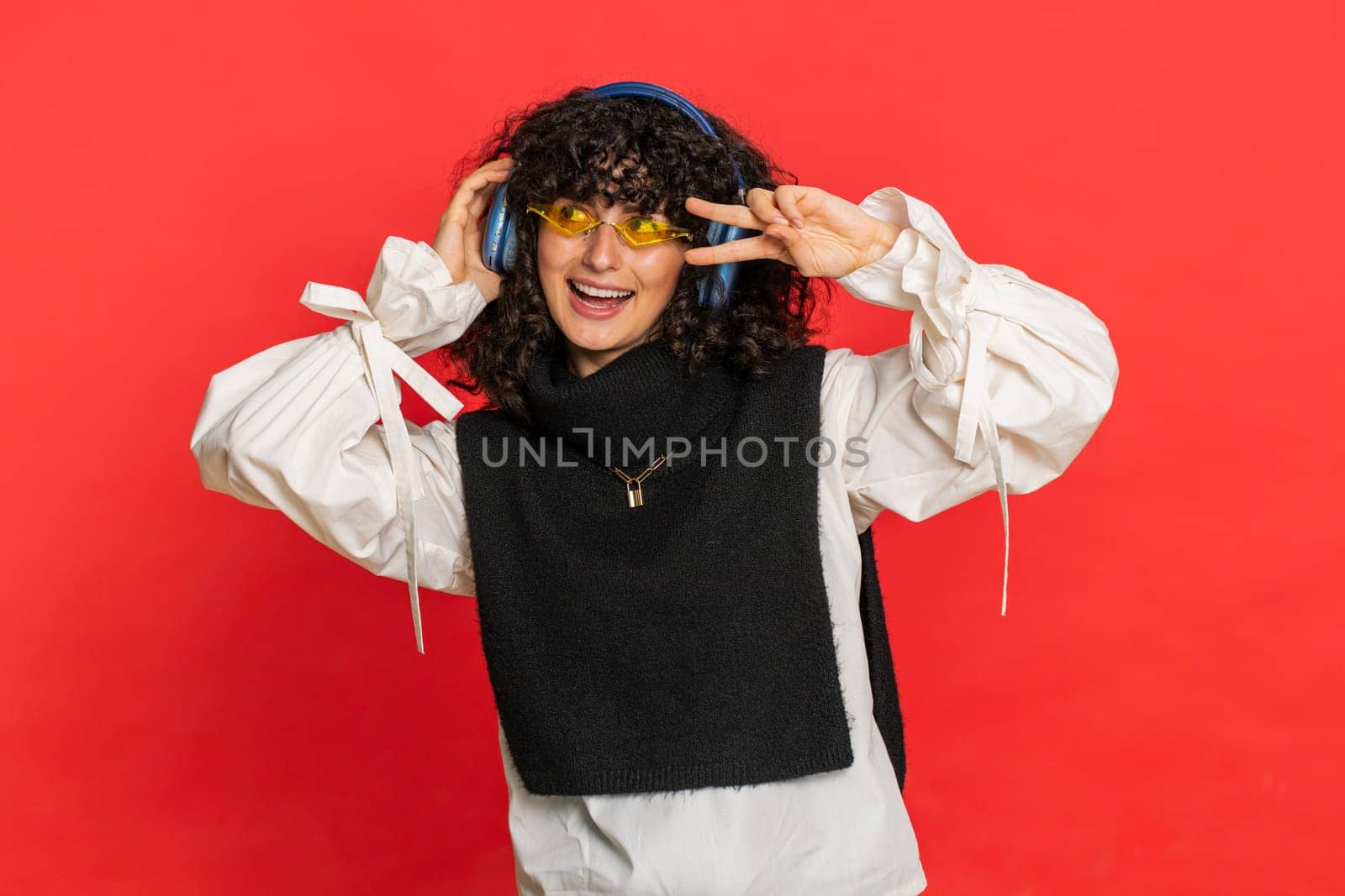 Caucasian young woman with curly hair listening music on headphones, dancing disco fooling around having fun expressive gesticulating hands. Music app advertisement. Girl isolated on red background