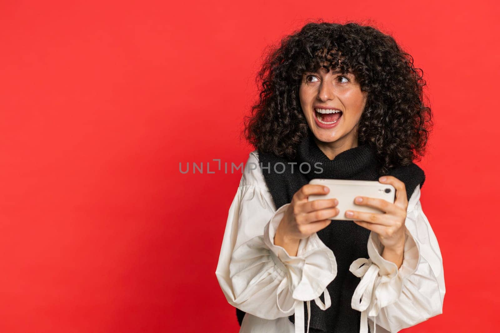 Happy excited Caucasian young woman with curly hair playing racing drive simulator shooter video games on smartphone. Girl player celebrate victory lottery jackpot goal achievement on red background