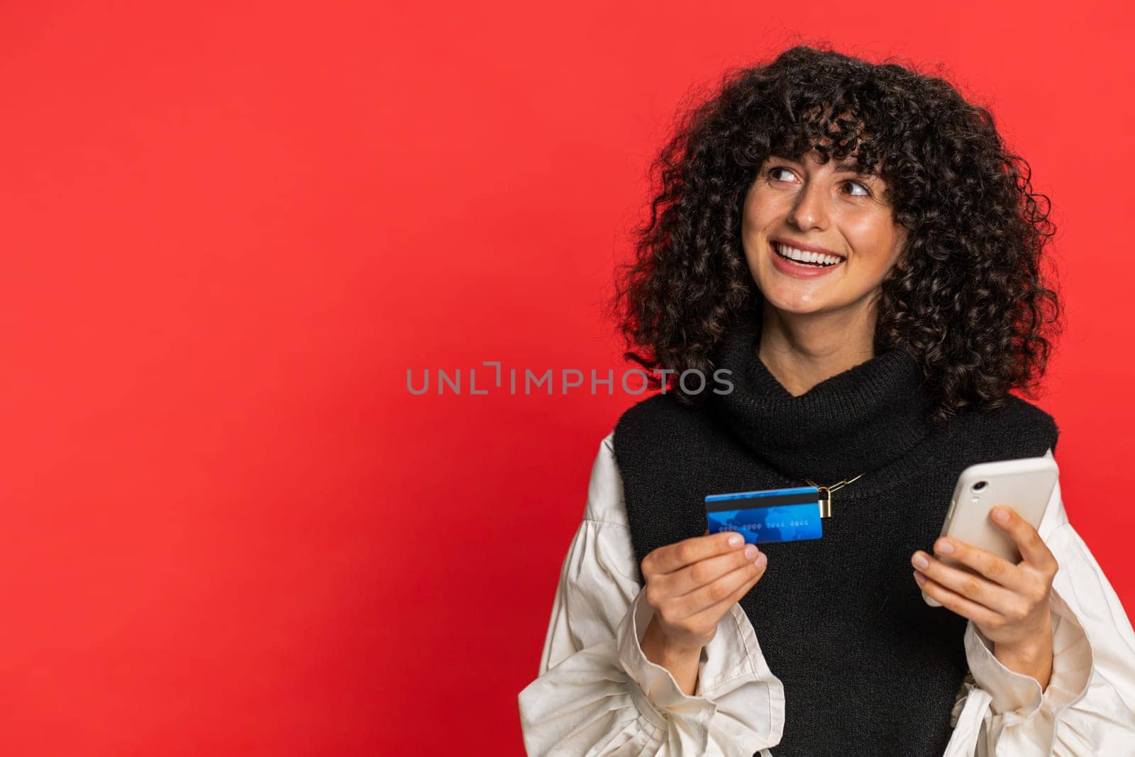 Happy Caucasian young woman with curly hair using credit bank card and smartphone while transferring spending money, purchases online shopping, ordering food delivery. Girl isolated on red background