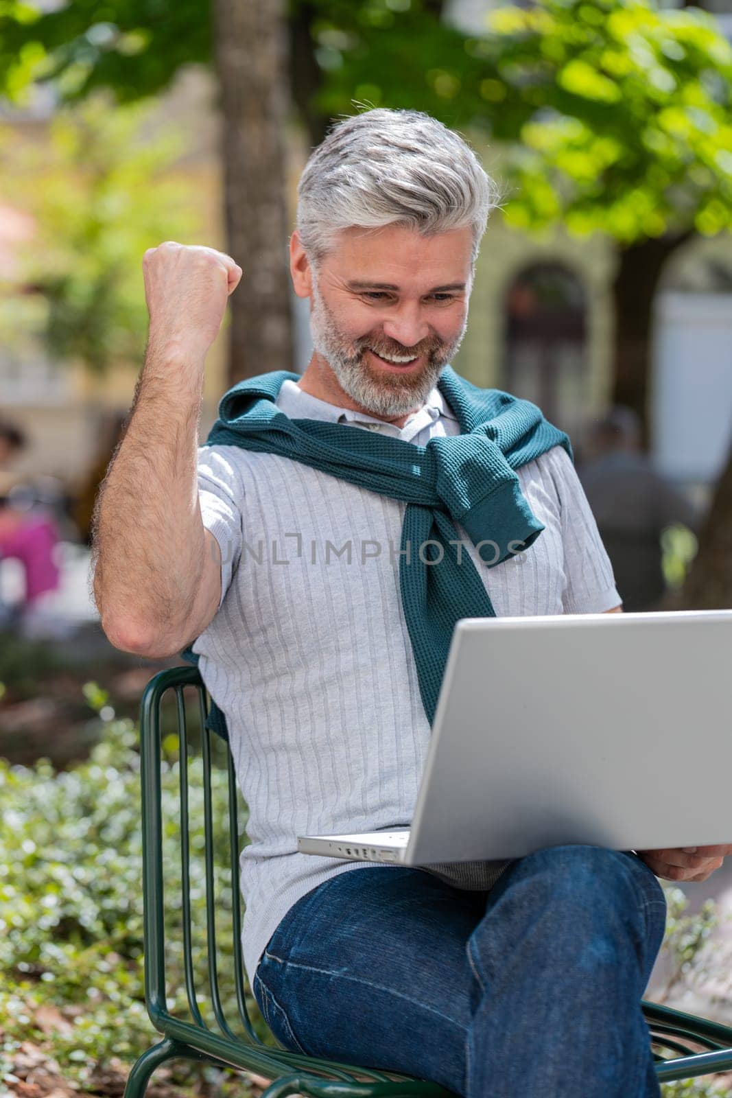 Handsome mature man using laptop computer celebrating win good message news, lottery jackpot victory, giveaway online outdoors. Happy bearded guy tourist sits in city cafe terrace. Town lifestyles.