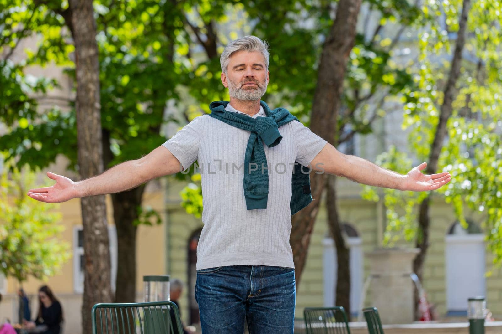 Keep calm down, relax. Bearded mature man meditating, breathes deeply with mudra gesture, praying eyes closed, concentrated thoughts, peaceful mind outdoors. Guy taking a break in urban city street.