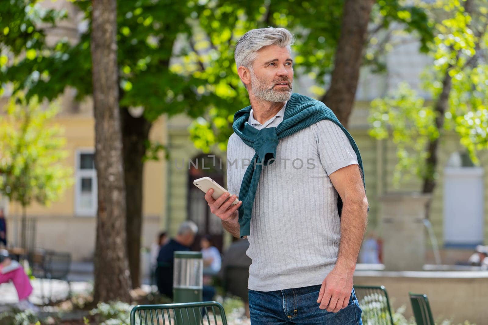 Tourist traveler mature man using smartphone text messages looking for search a way on map in mobile navigator app outdoors. Caucasian bearded guy male walking passes by urban city street background.