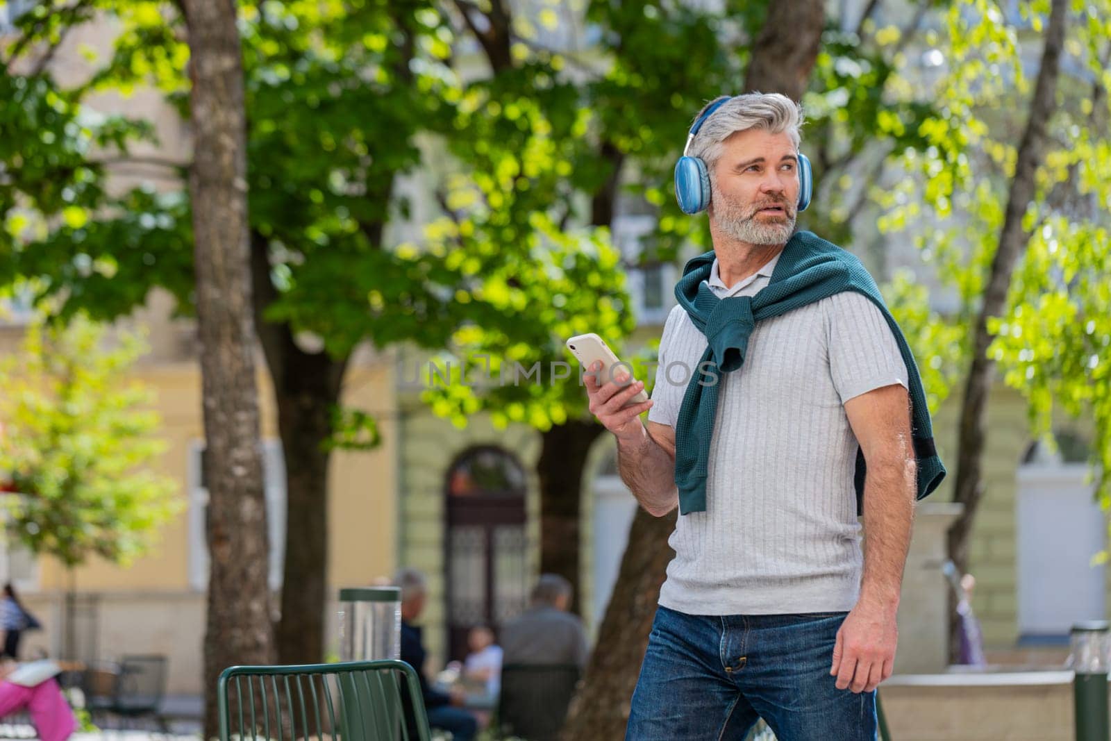 Happy mature Caucasian man in wireless headphones choosing, listening favorite energetic music in smartphone outdoors. Bearded guy tourist walking passes by urban sunshine city street. Lifestyles.