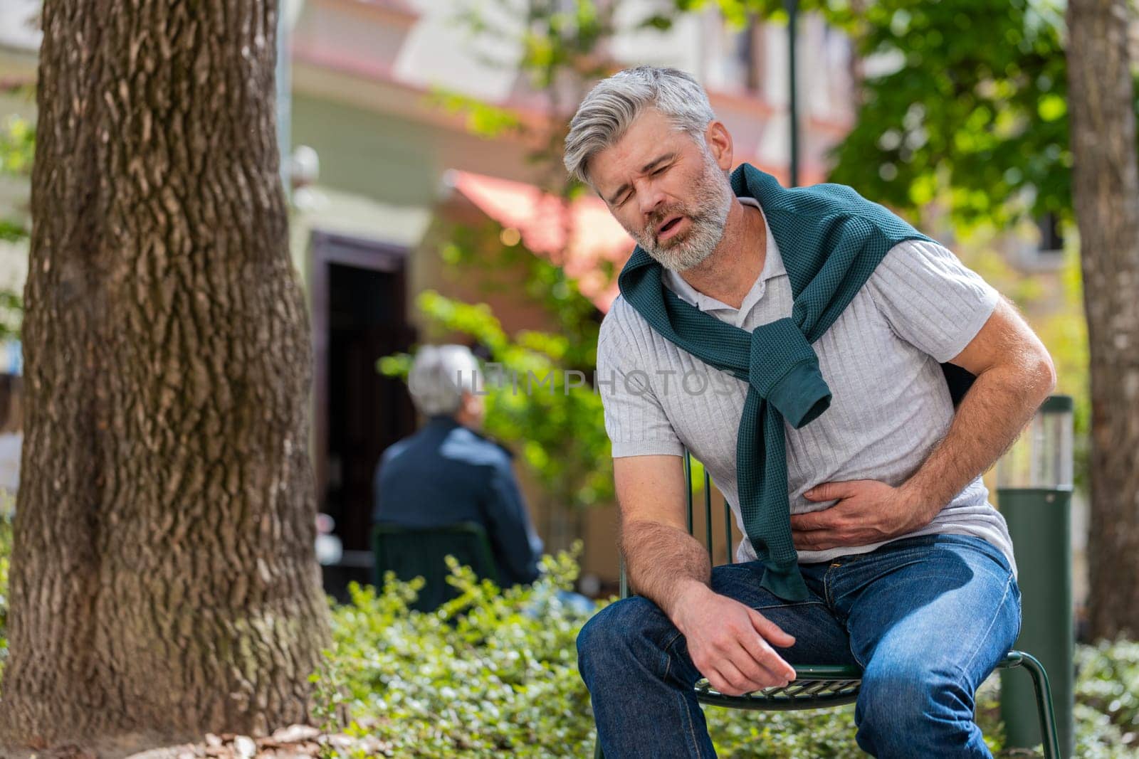 Caucasian mature man feeling sudden strong abdominal stomach ache, gastritis problem. Upset tourist guy having symptom poisoning diarrhea indigestion peptic ulcer pancreatitis on city urban street.