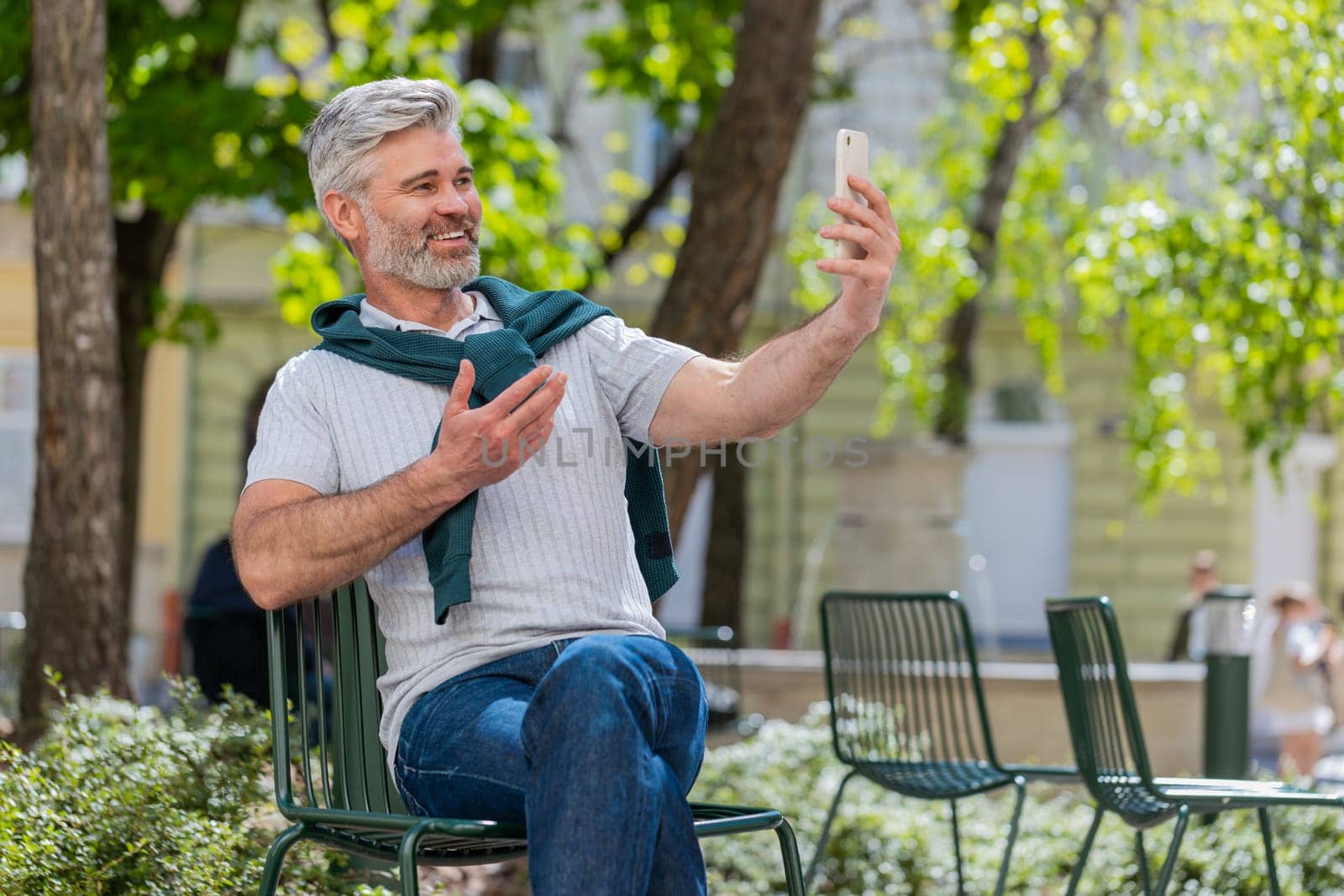 Bearded mature man blogger using smartphone, communicating video call online with subscribers, recording stories for social media vlog outdoors. Guy sitting on chair in urban sunshine city street.