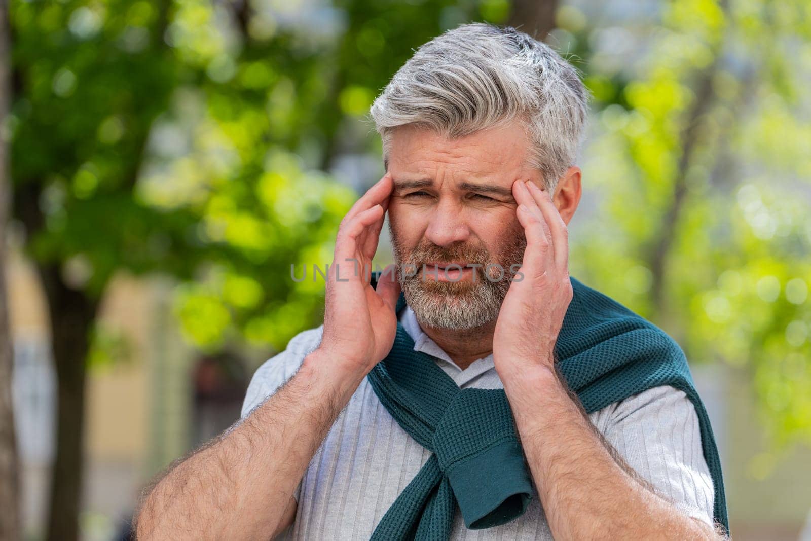 Displeased exhausted Caucasian man rubbing temples to cure headache problem suffering from tension migraine stress. Mature bearded guy tourist grimacing, pain standing on city urban street town.