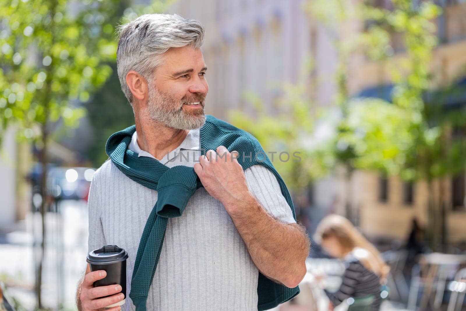 Happy Caucasian bearded man enjoying morning coffee hot drink and smiling. Relaxing, taking a break. Mature guy walking on urban city center street, drinking coffee to go. Town lifestyles outside.