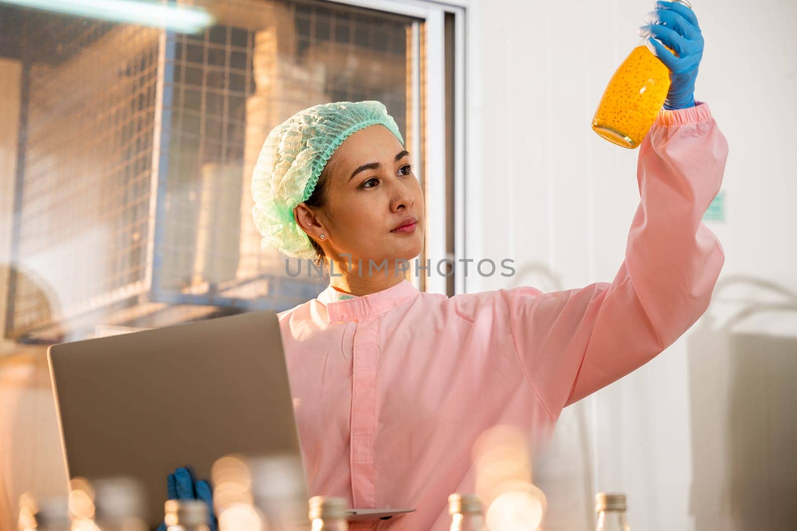 In a beverage factory a woman part of quality control meticulously inspects bottles on a conveyor belt. Using a laptop she ensures liquid quality maintaining industry standards.