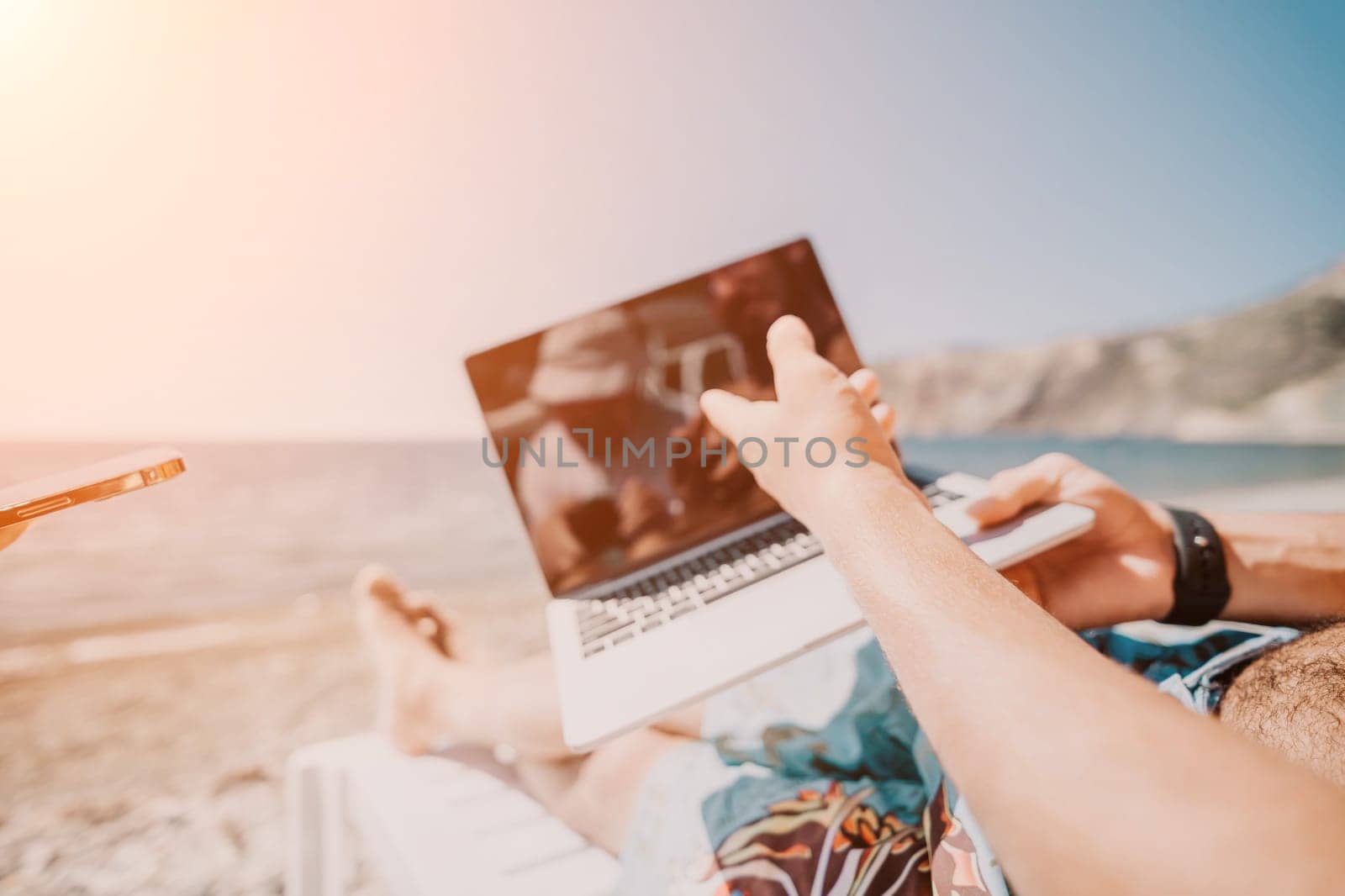 Digital nomad, Business man working on laptop by the sea. Man typing on computer by the sea at sunset, makes a business transaction online from a distance. Freelance, remote work on vacation