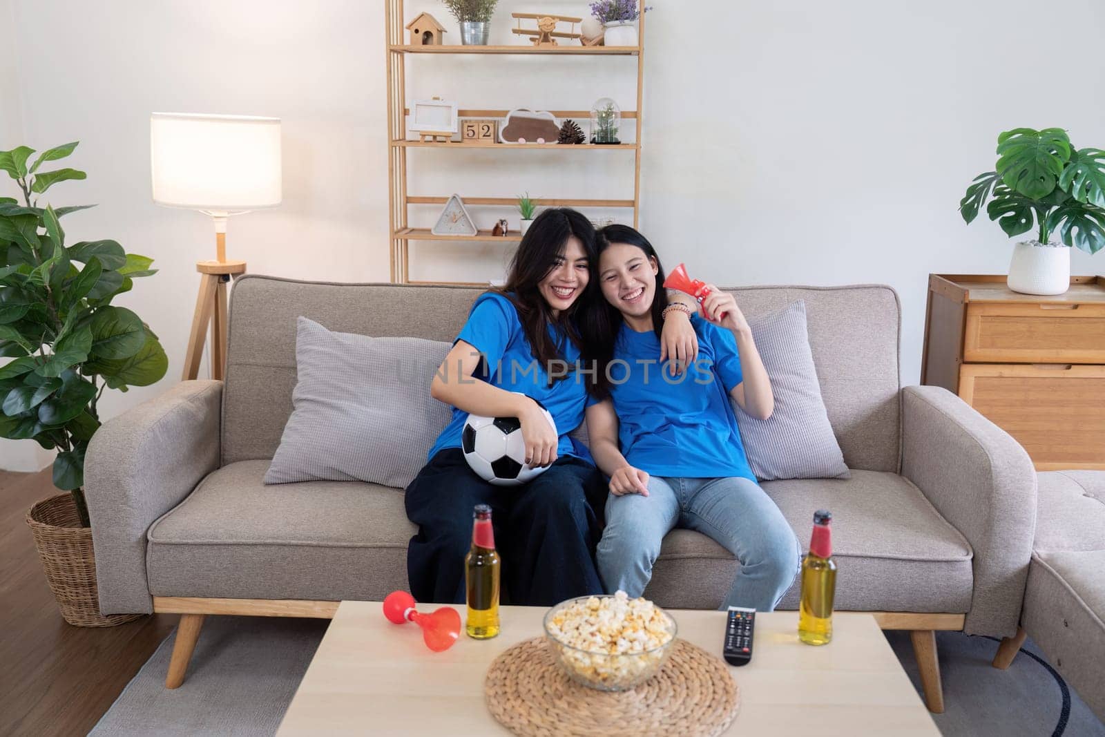 Lesbian couple cheering for Euro football match at home with drinks and popcorn. Concept of LGBTQ pride, sports enthusiasm, and celebration.