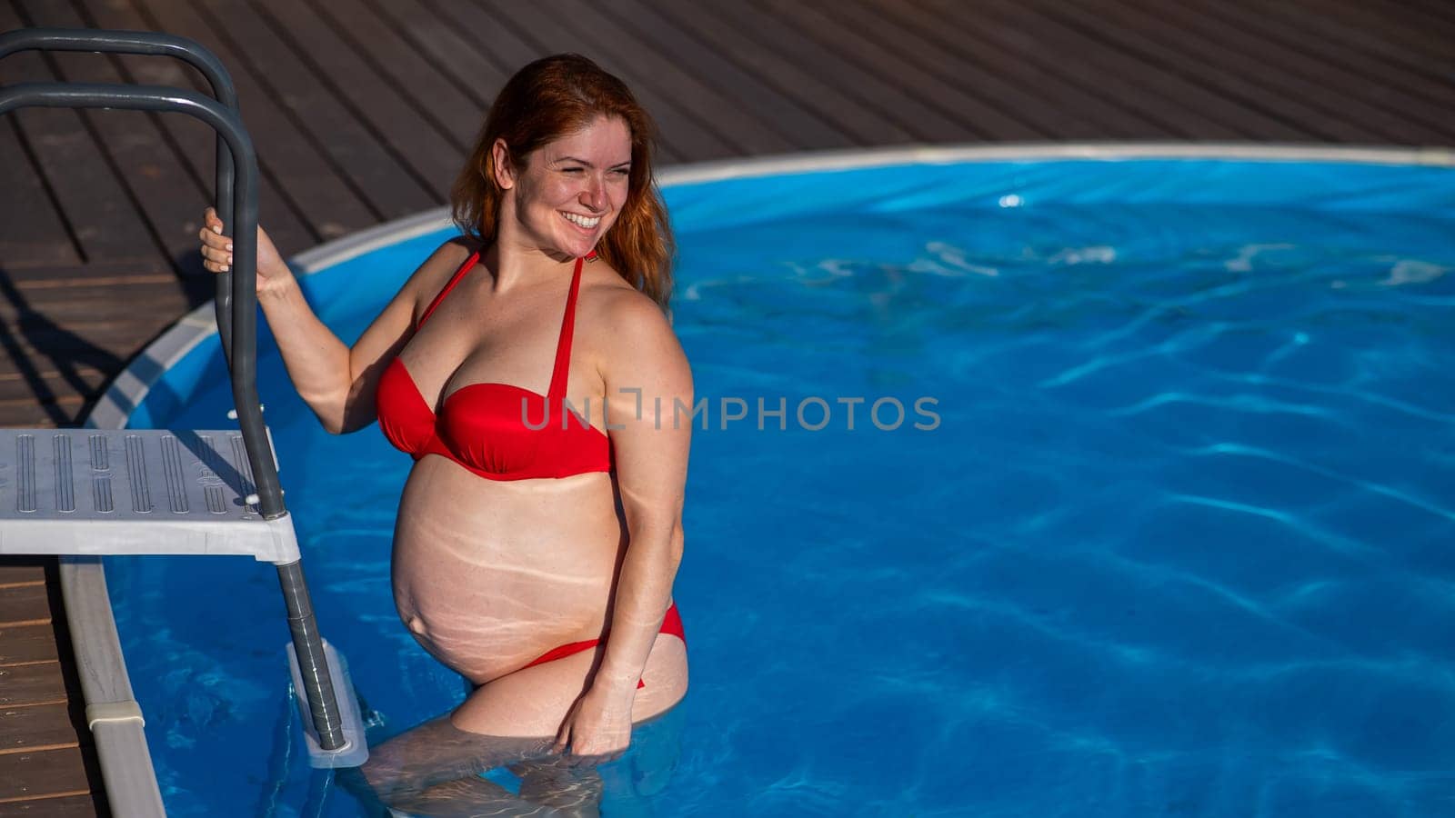 A red-haired pregnant woman goes down the stairs into the pool outdoors
