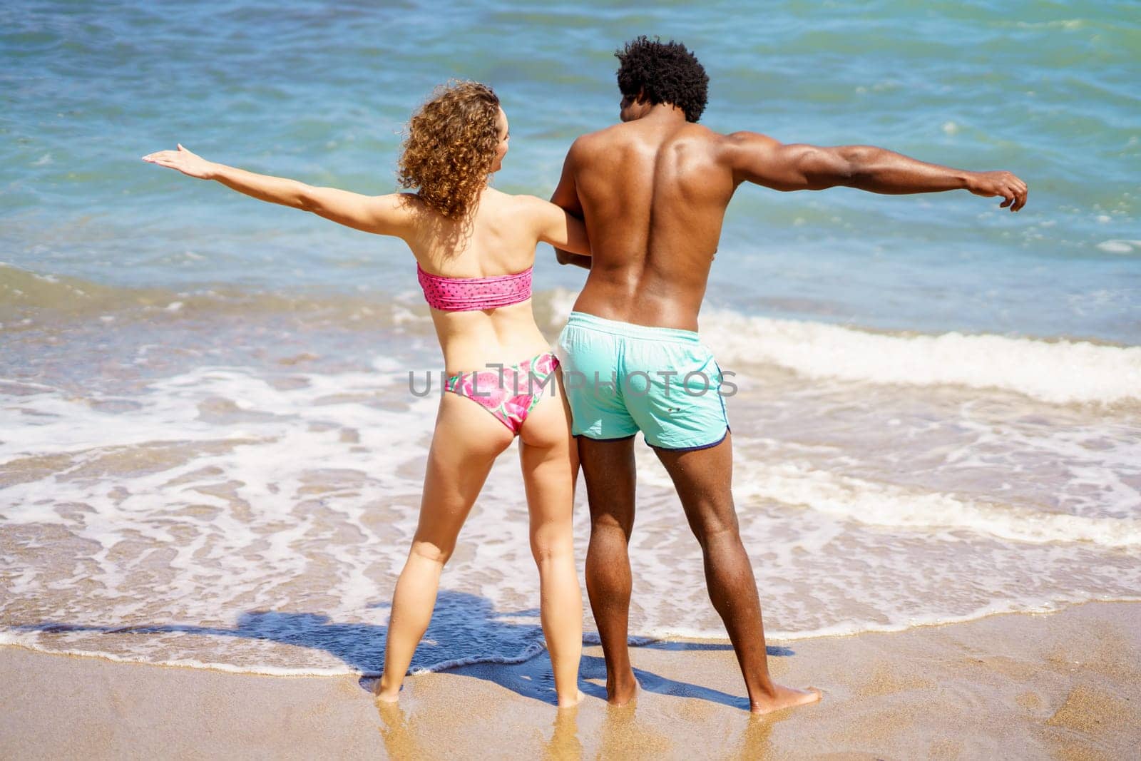 Full body back view of anonymous African American boyfriend and curly haired girlfriend in swimwear having fun near foamy sea during summer journey