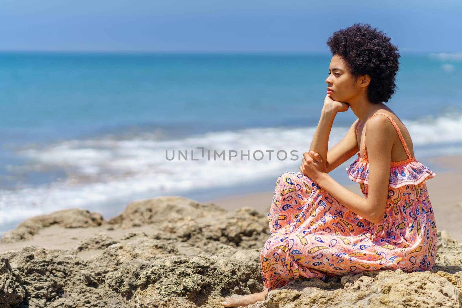 Pensive young African American female tourist, in maxi dress with curly hair, leaning on hand and looking at waving sea while sitting on rough stone on summer day on beach
