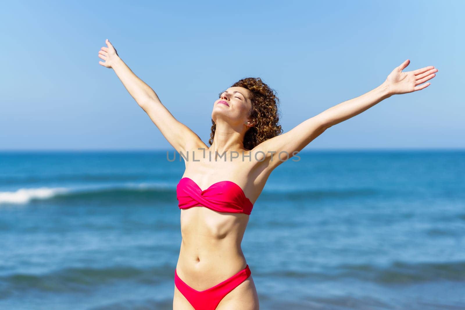 Content female with closed eyes in pink swimsuit raising arms and smiling while sunbathing and resting against blue sky and waving sea on summer holiday