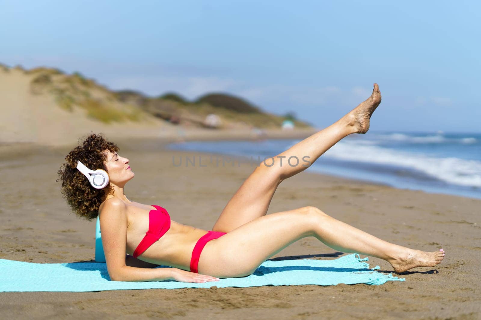 Full body side view of young slim female in pink bikini and headphones raising leg while lying on sandy beach under sunshine
