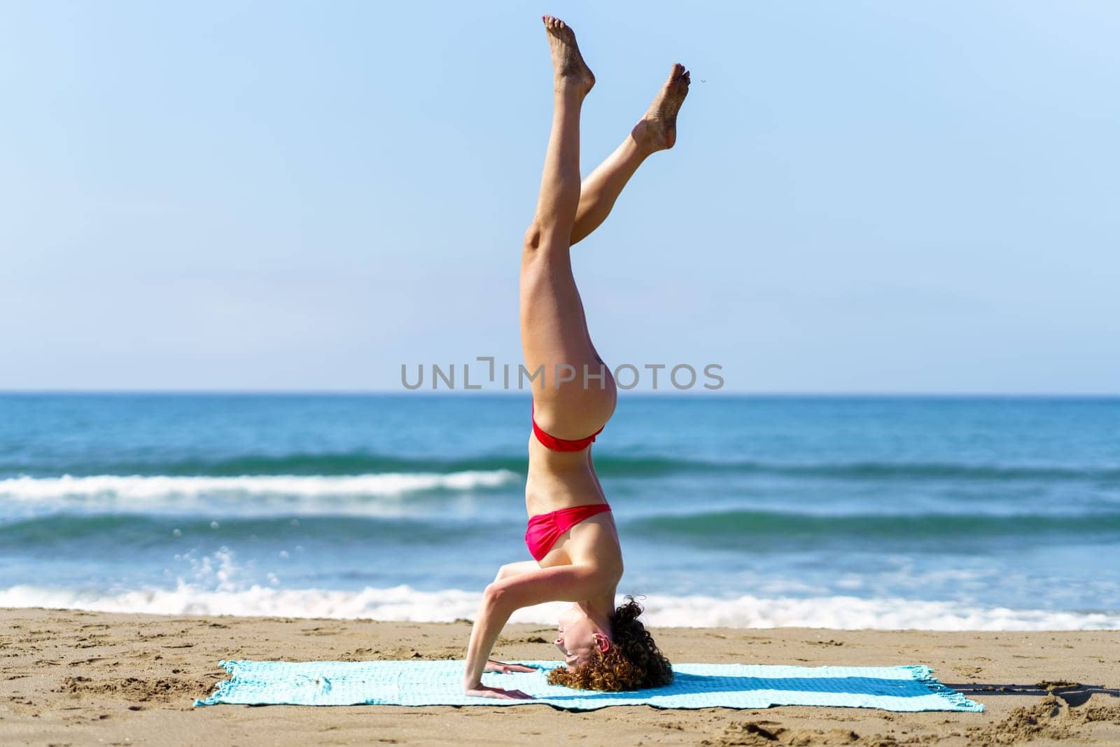 Side view of young redhead lady in bikini doing Tripod Headstand on coastline of ocean with blue sky