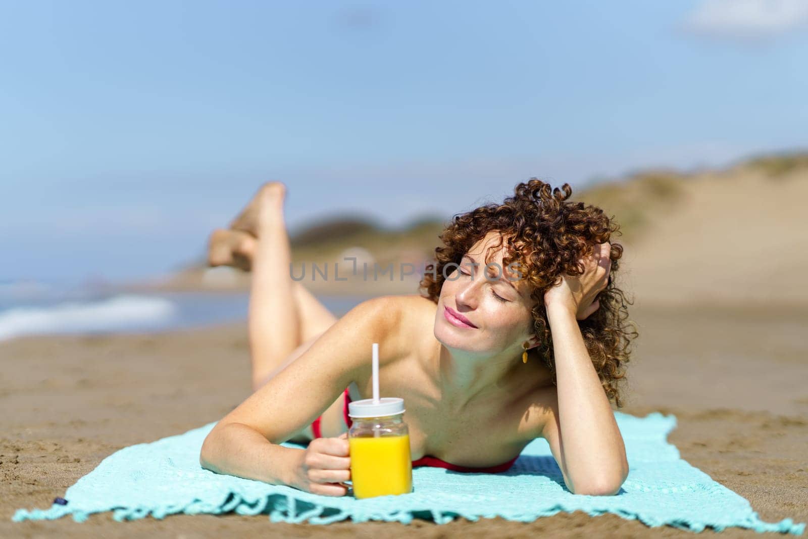 Cheerful young woman with curly hair and closed eyes lying on sandy beach with glass of orange juice on sunny day