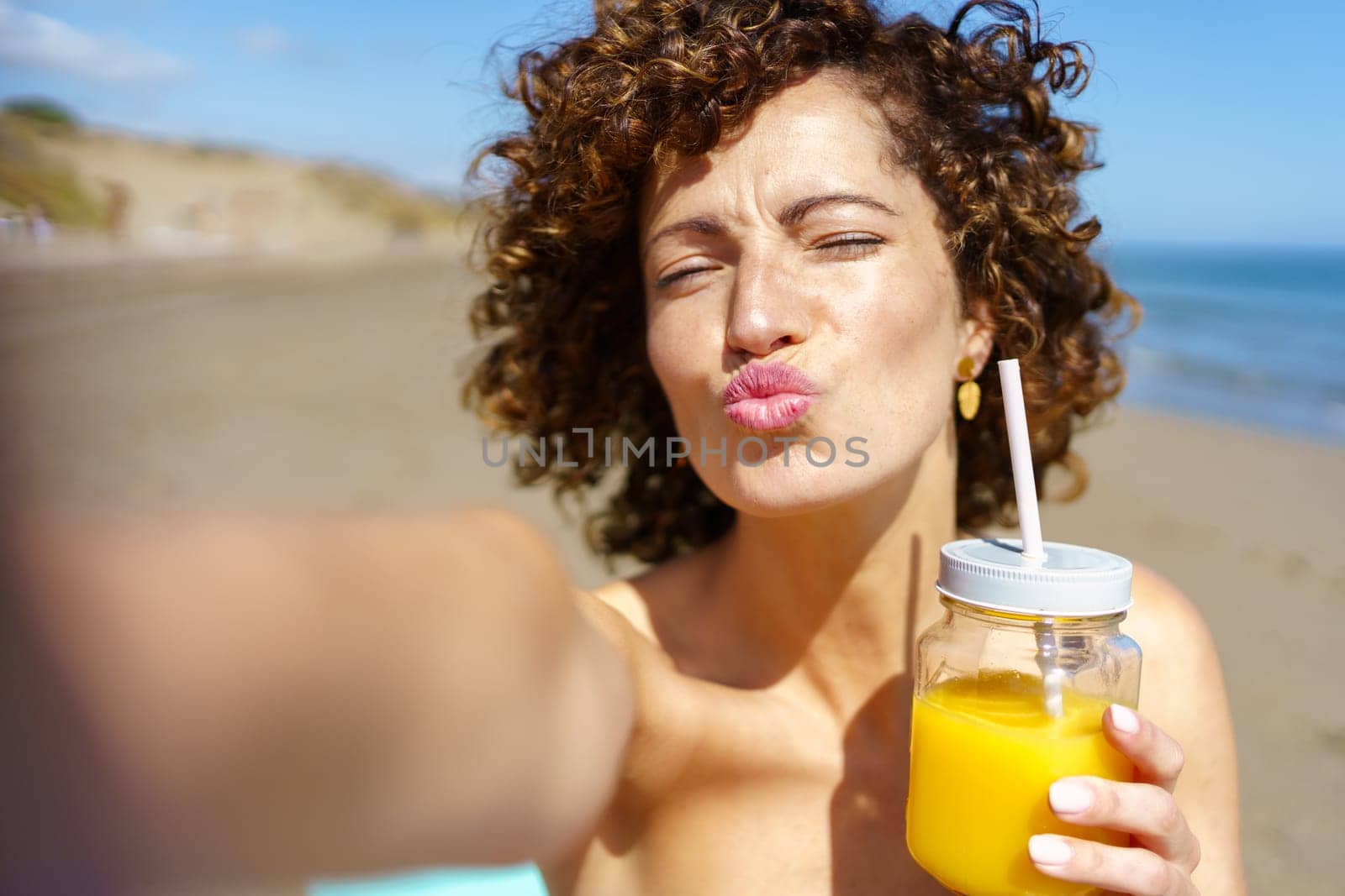 Young female with curly hair and closed eyes blowing kiss and holding jar of orange juice while taking selfie on beach during summer vacation