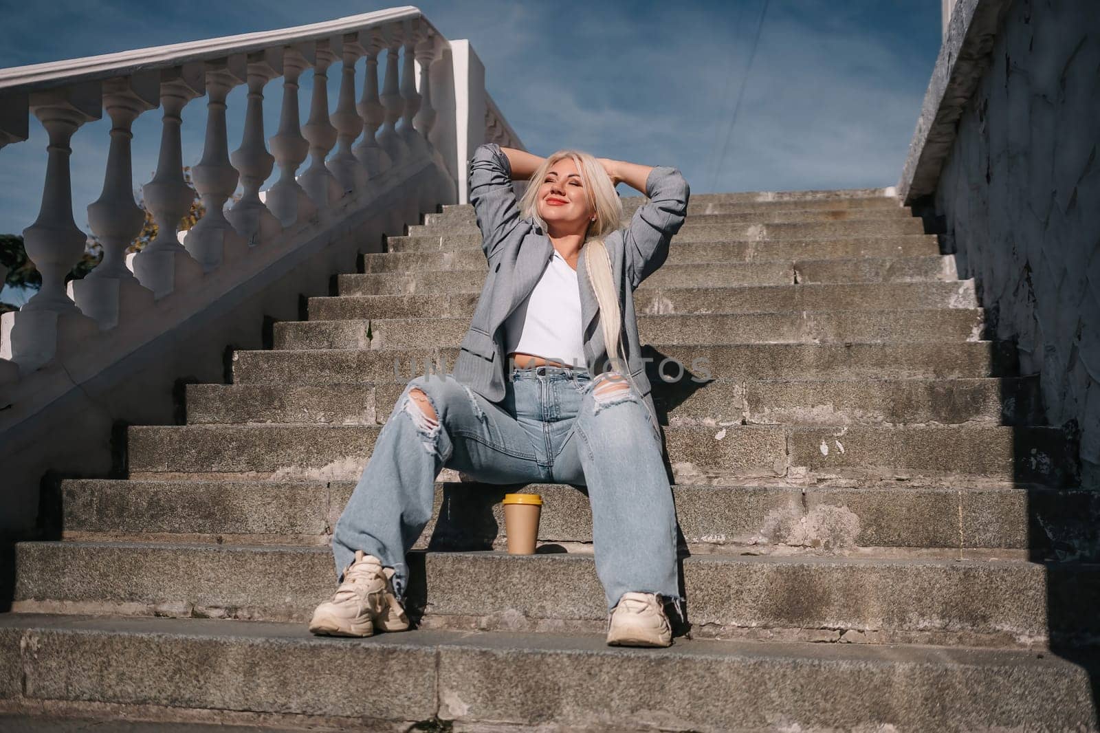 A woman in jeans and a jacket sits on a set of stairs. She is smiling and holding a cup
