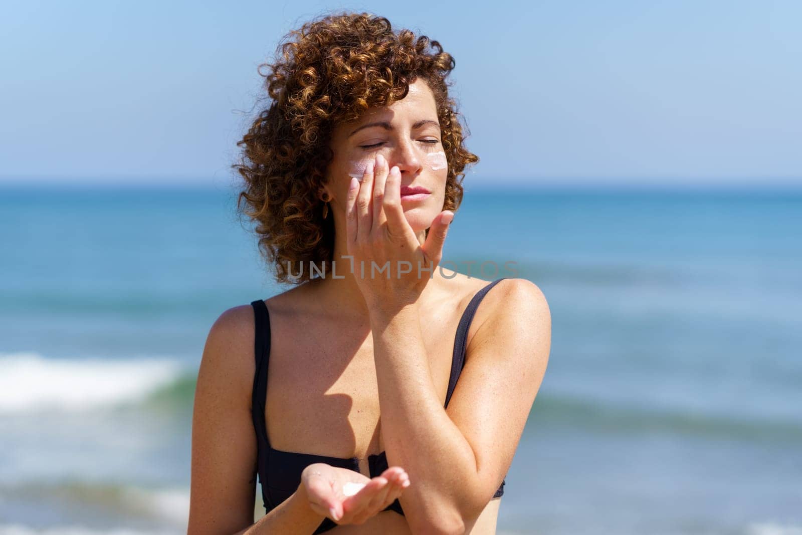 Sensual woman in bikini standing near waving ocean while applying sunblock cream on face during summer vacation