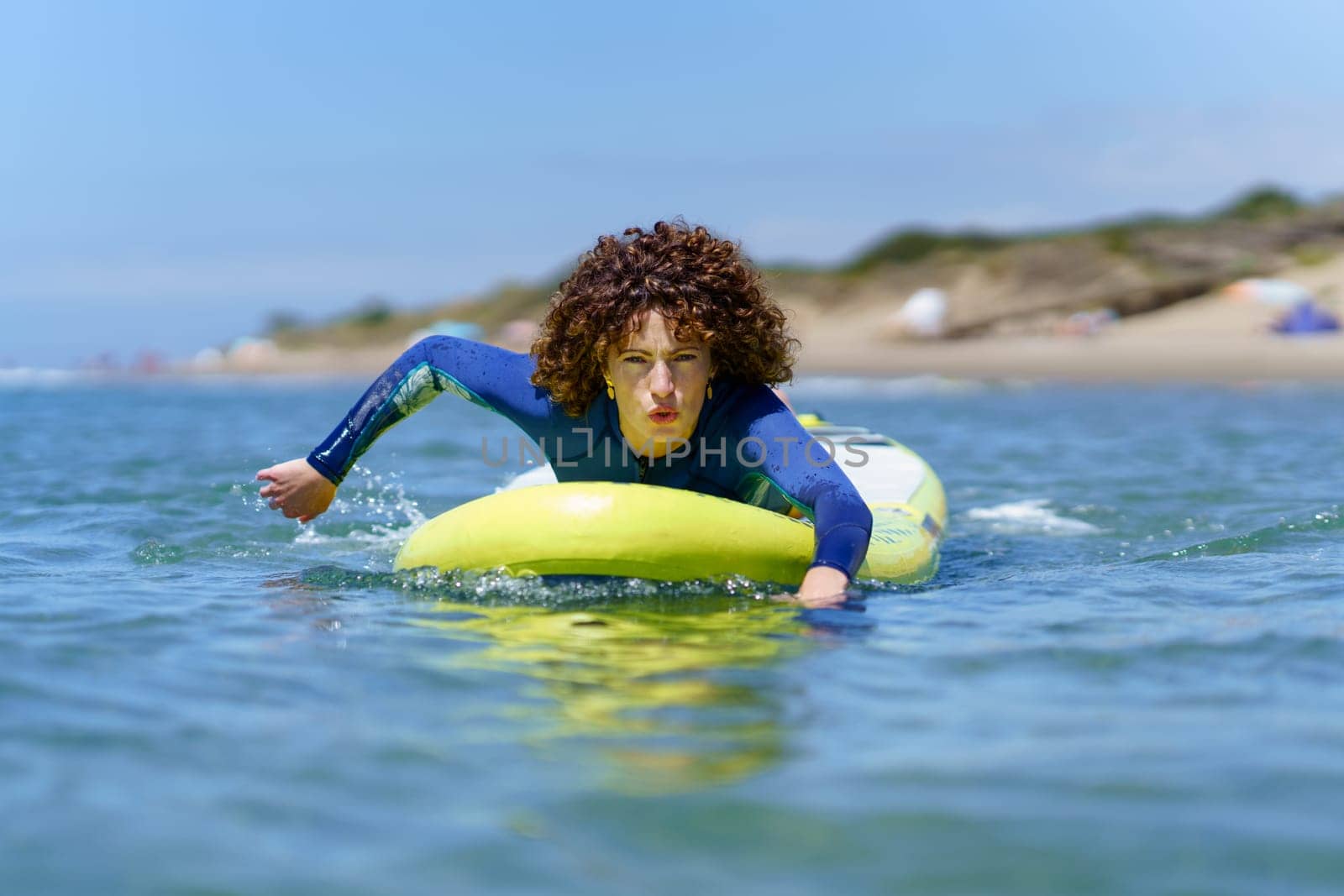 Concentrated young female surfer in wetsuit with curly hair lying on SUP board while paddling with hands floating on ocean during summer vacation