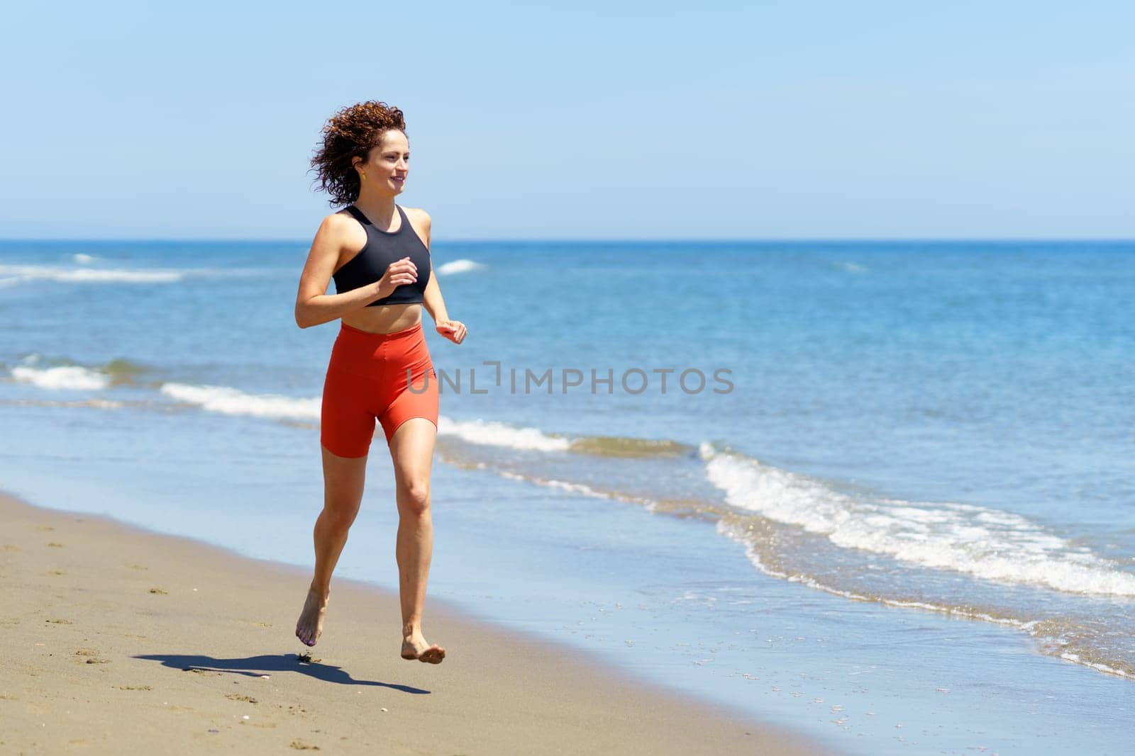 Side view of smiling barefoot young fit female athlete, in sportswear warming up and looking away while jogging on sandy beach near waving sea against cloudless blue sky