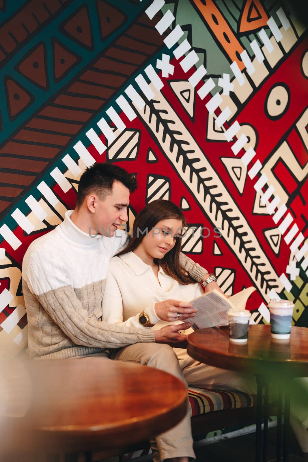 A man and a woman are seated at a wooden table, focused on reading a book. The man is wearing a blue shirt and glasses, while the woman has long hair and a pink sweater. The table is cluttered with books and a cup of coffee.