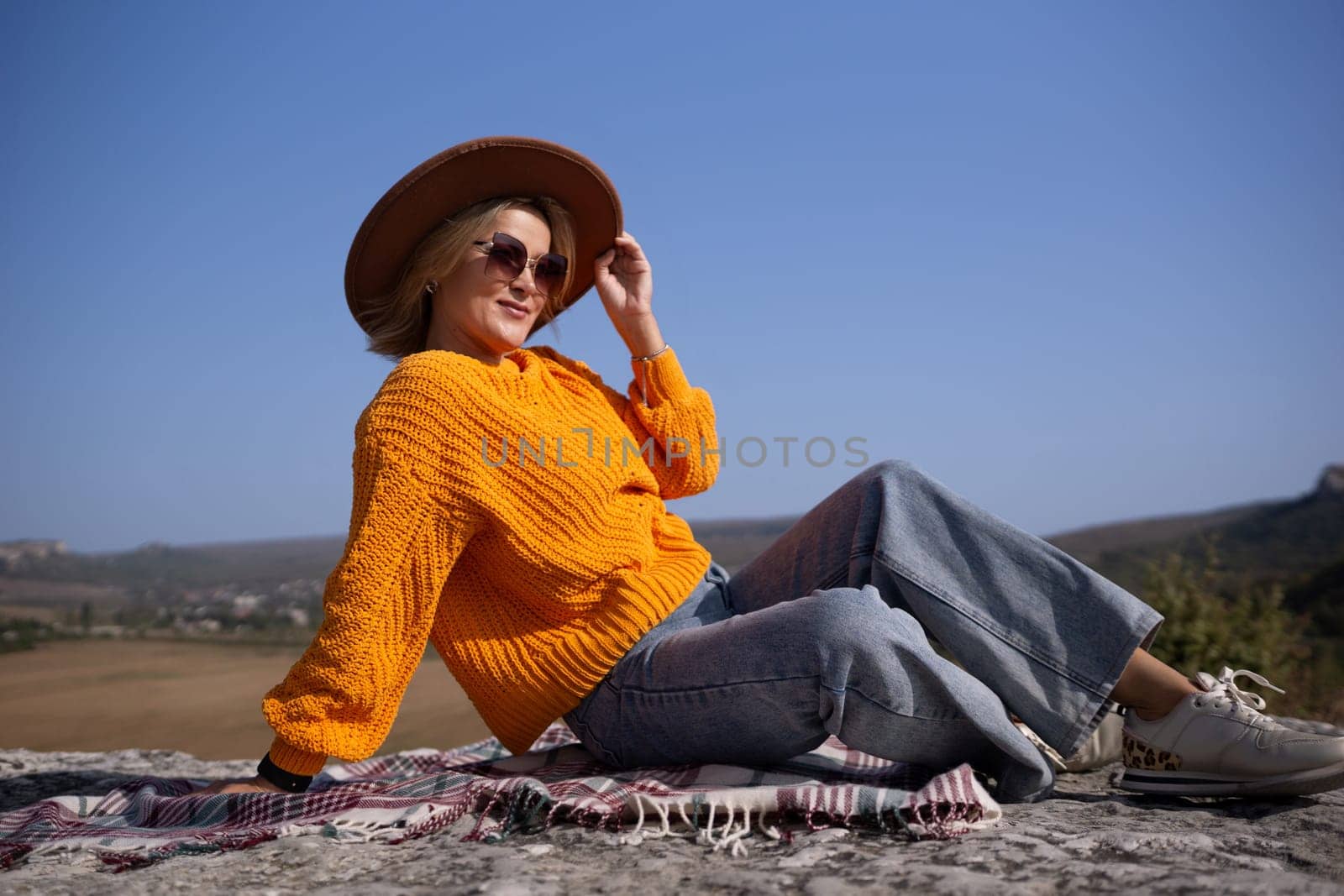 A woman in a yellow sweater and blue jeans is sitting on a blanket on a hillside. She is wearing sunglasses and a brown hat