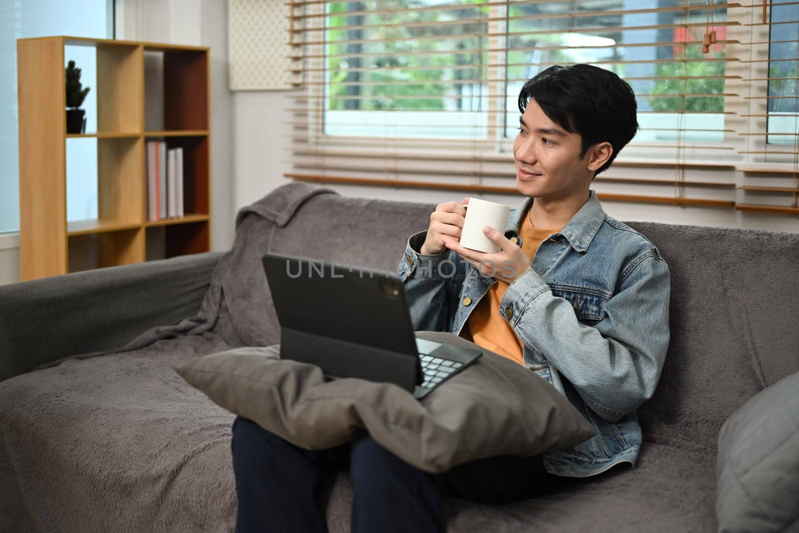 Satisfied male freelancer sitting on couch with digital tablet drinking coffee and looking away.