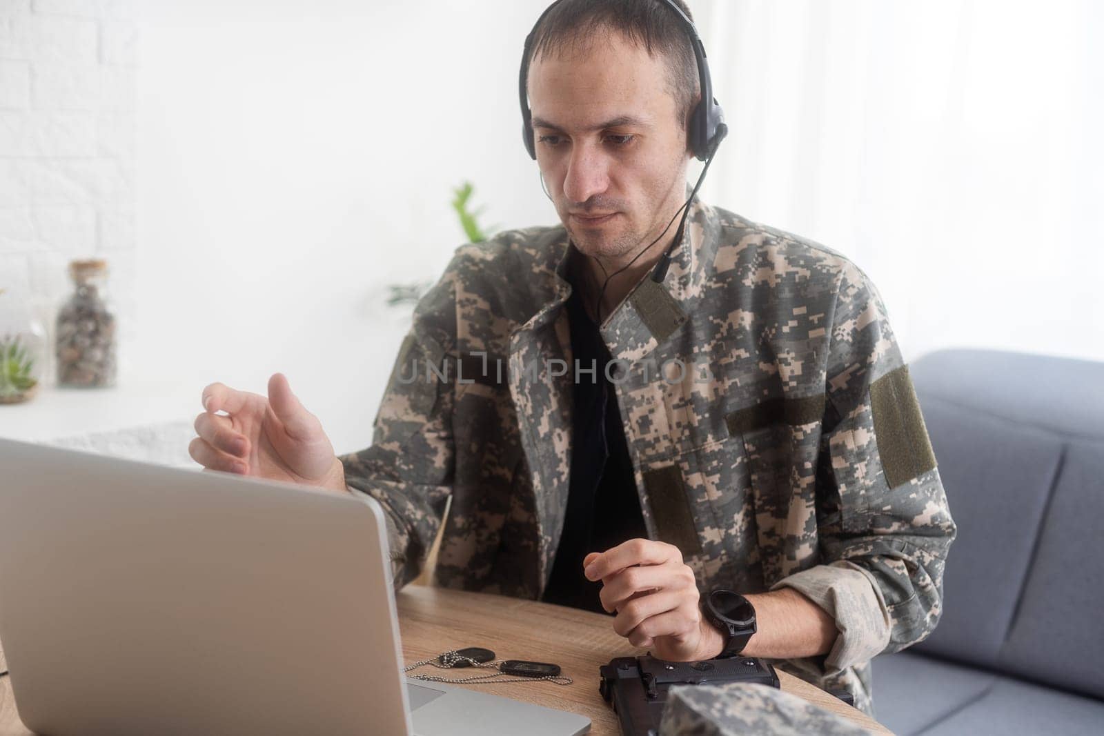 Soldier in office. Serious young military man typing on laptop while working in headquarters building. Caucasian man in camouflage uniform is sitting in bright room at table with notebook and cap. High quality photo