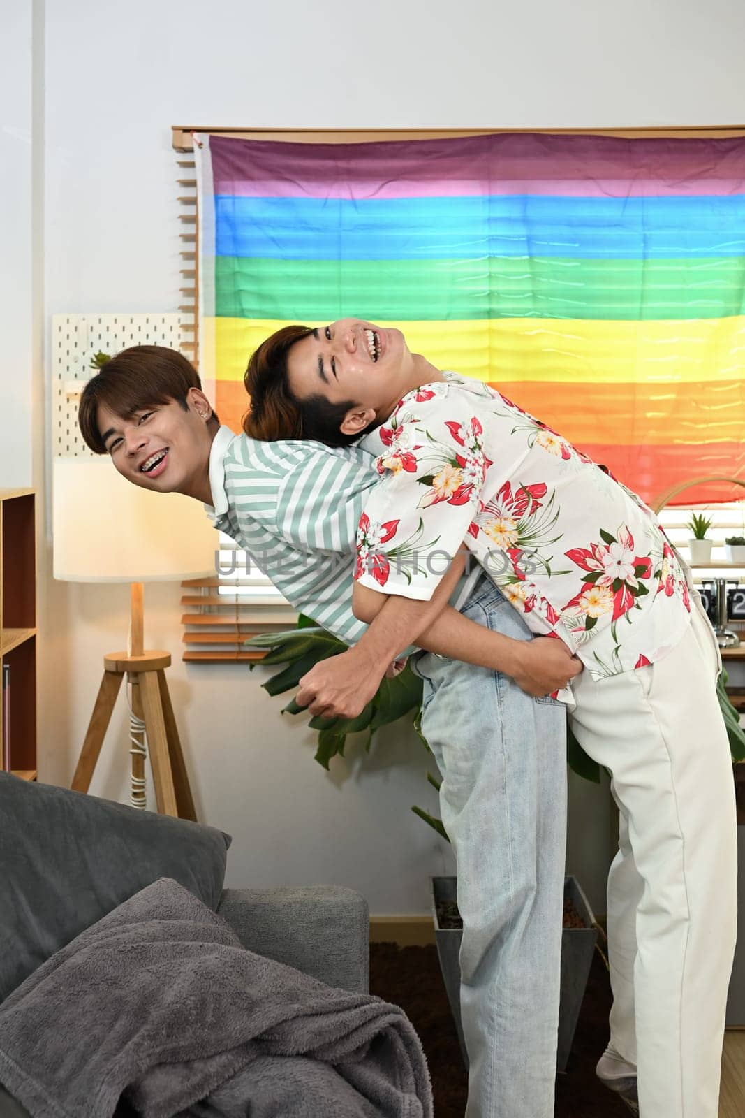 Cheerful gay male couple giving piggyback ride for each other laugh joyfully in living room.