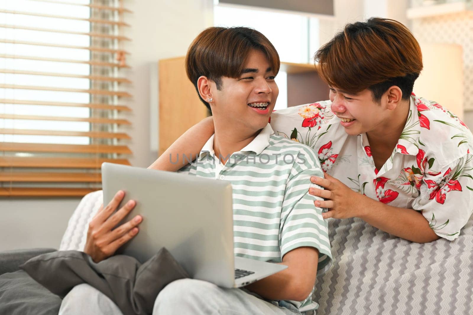 Cheerful gay couple looking at each other with tender and browsing laptop on couch at home.