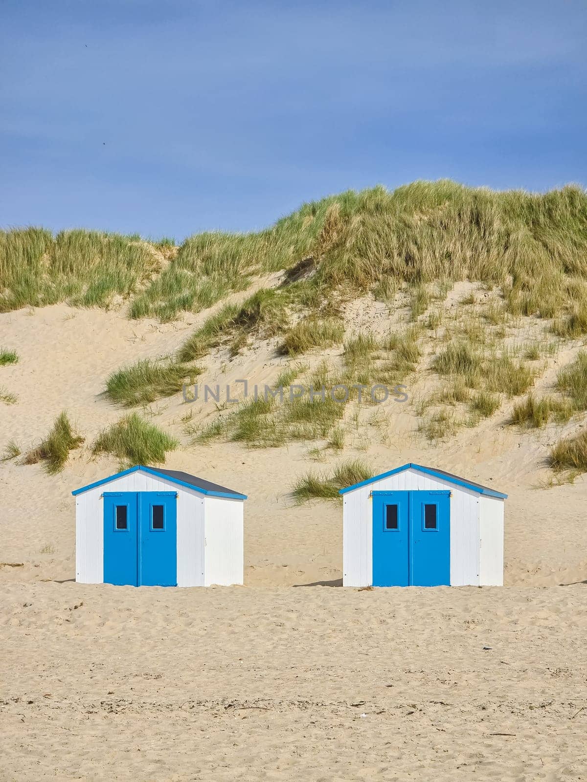 Three charming beach huts with blue doors stand on the sandy shores of Texel, Netherlands, against a backdrop of serene seaside beauty.