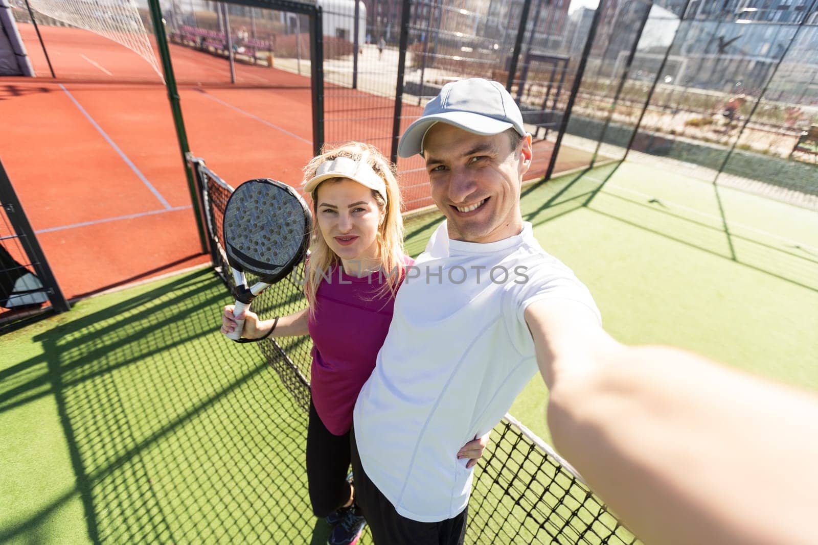 Young couple of tennis players wearing a sportswear holding a racket on their shoulders and a ball in a hand standing outdoor on tennis court at early morning. High quality photo