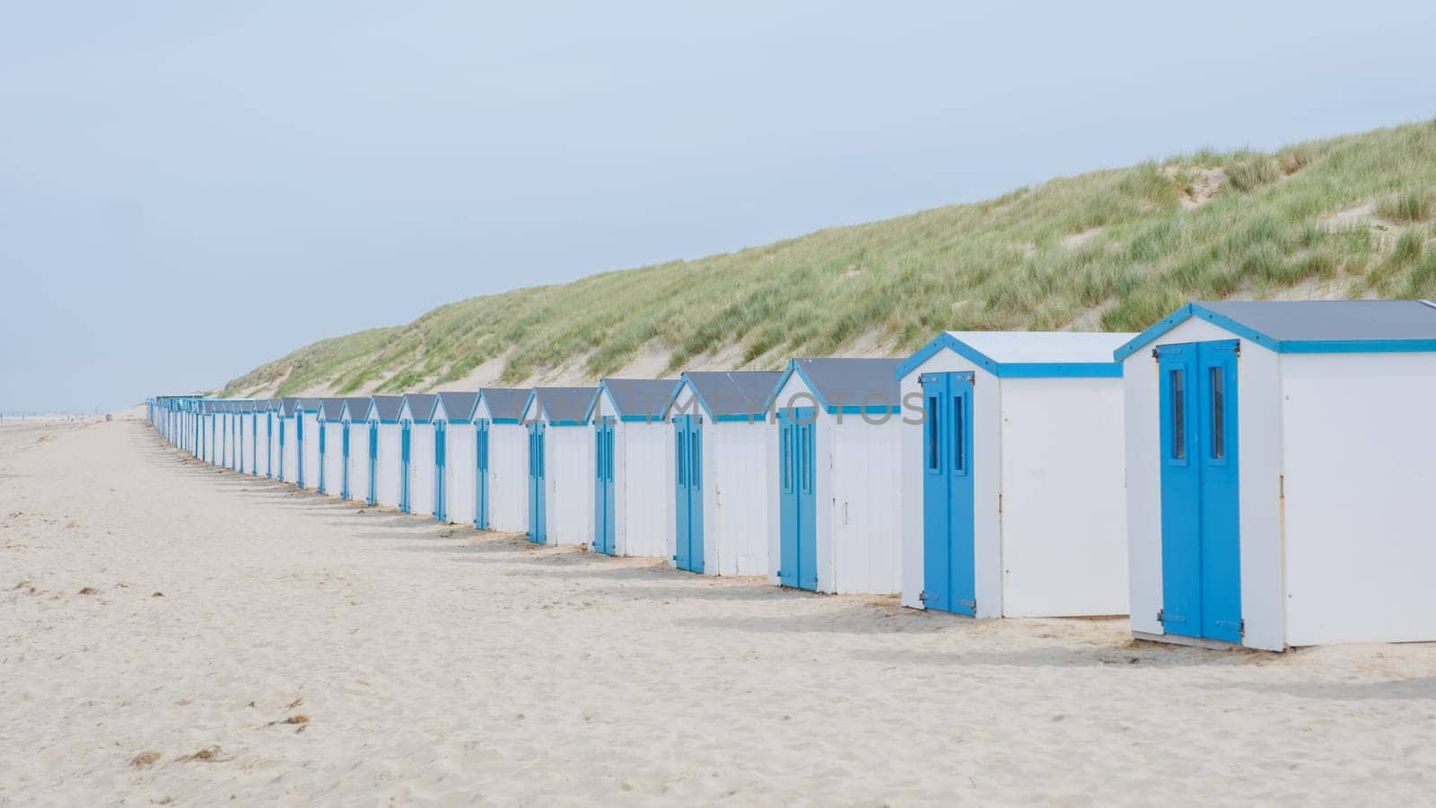 A picturesque row of blue and white beach huts stand elegantly on the sandy shores of Texel, Netherlands, under the clear blue sky.