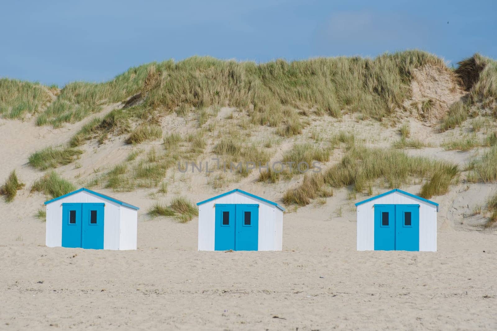 Beach huts painted in a rainbow of colors line the sandy shore on Texel, Netherlands, under a clear blue sky.