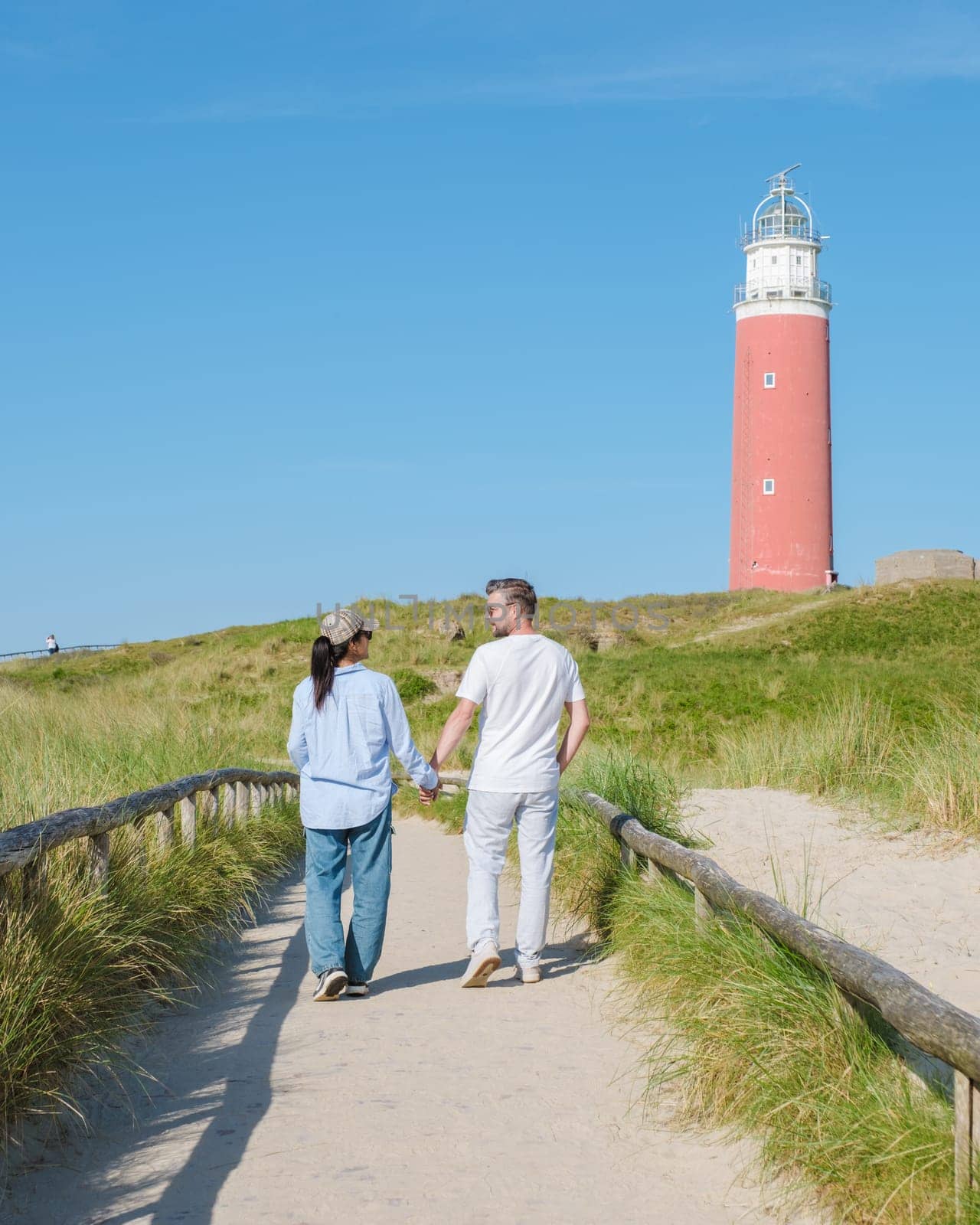 A couple strolls hand in hand on a winding path, gazing out at the iconic lighthouse along the Texel, Netherlands shoreline. The iconic red lighthouse of Texel Netherlands
