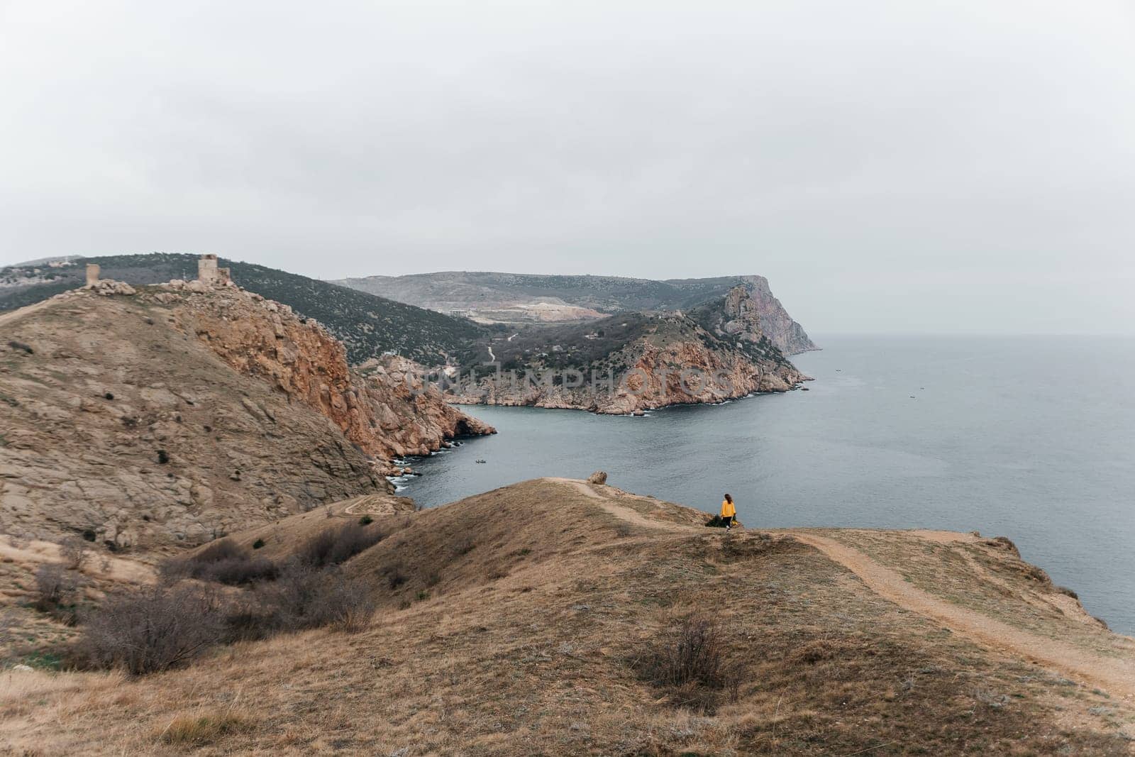Woman stands on cliff overlooking ocean, cloudy day provides soft lighting. She's wearing a yellow sweater wearing dark pants. Landscape features rugged terrain, calm waters