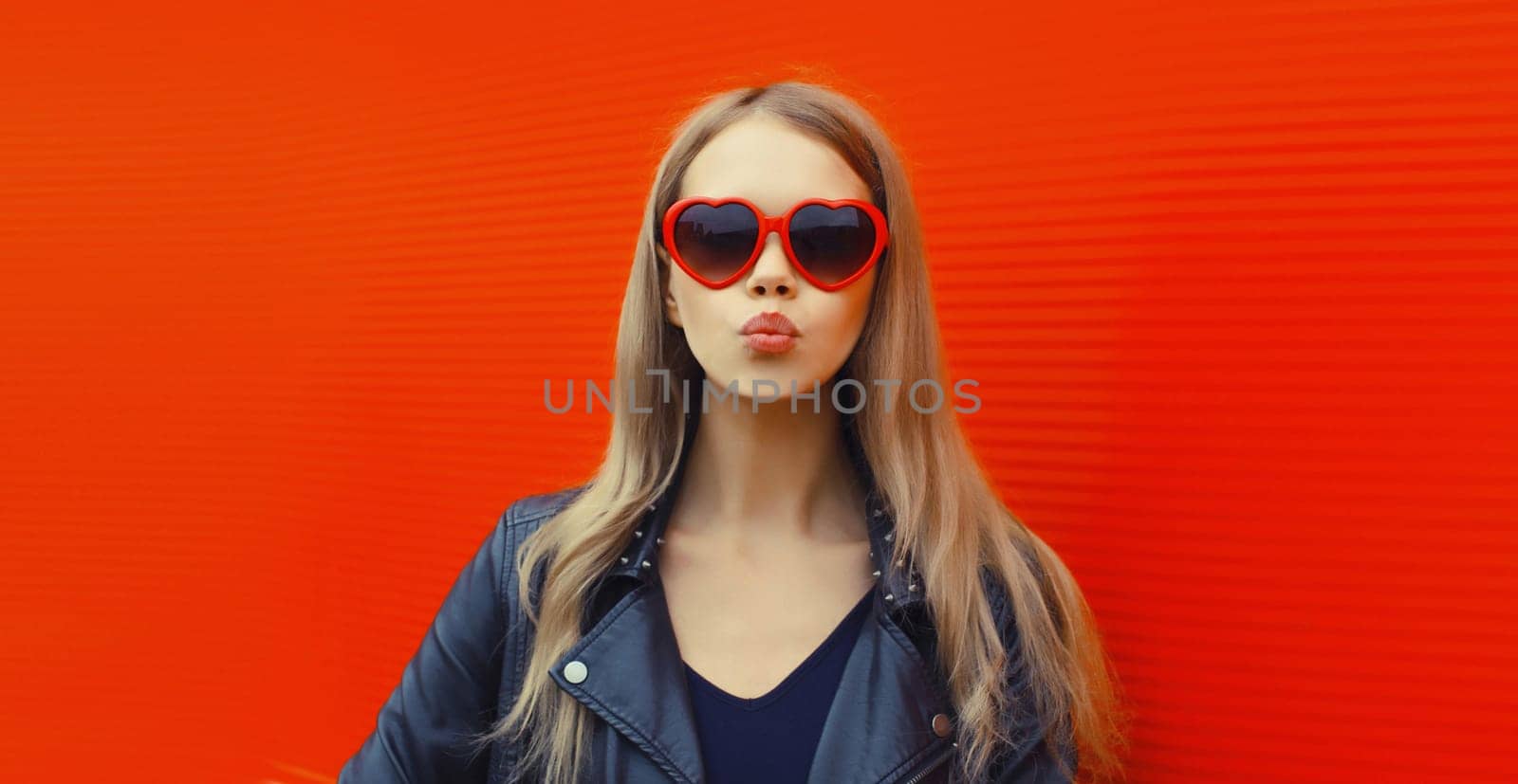 Portrait of beautiful stylish blonde young woman model blowing kiss in red heart shaped sunglasses, black rock leather jacket on city street