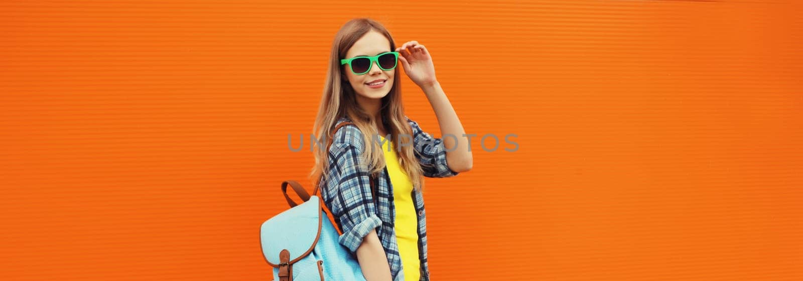 Summer portrait of happy cheerful stylish young woman with skateboard, backpack in colorful clothes posing on orange background on city street