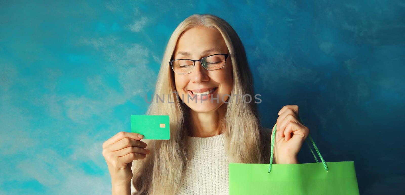 Portrait of happy smiling caucasian middle aged woman holding plastic credit bank card with shopping bags on blue background