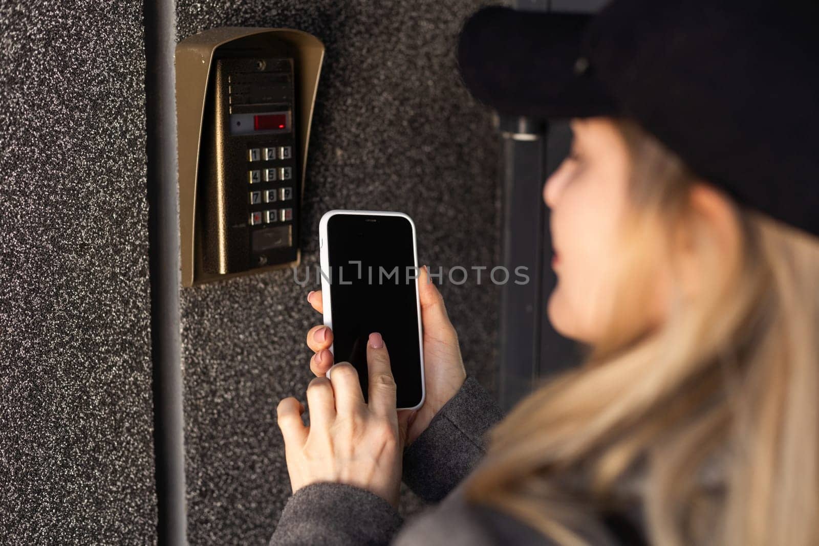Female hand holding black mobile smart phone with smart home application on the screen. Blurred house on the background. For access to all of the controls of your house and caring of home security. High quality photo