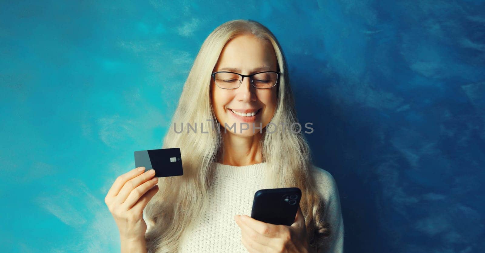 Portrait of happy smiling middle aged woman holding plastic credit bank card, looking at smartphone, shopping online or ordering goods
