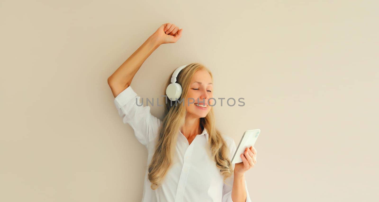 Happy cheerful middle aged woman listening to music with mobile phone in headphones having fun dancing on studio background