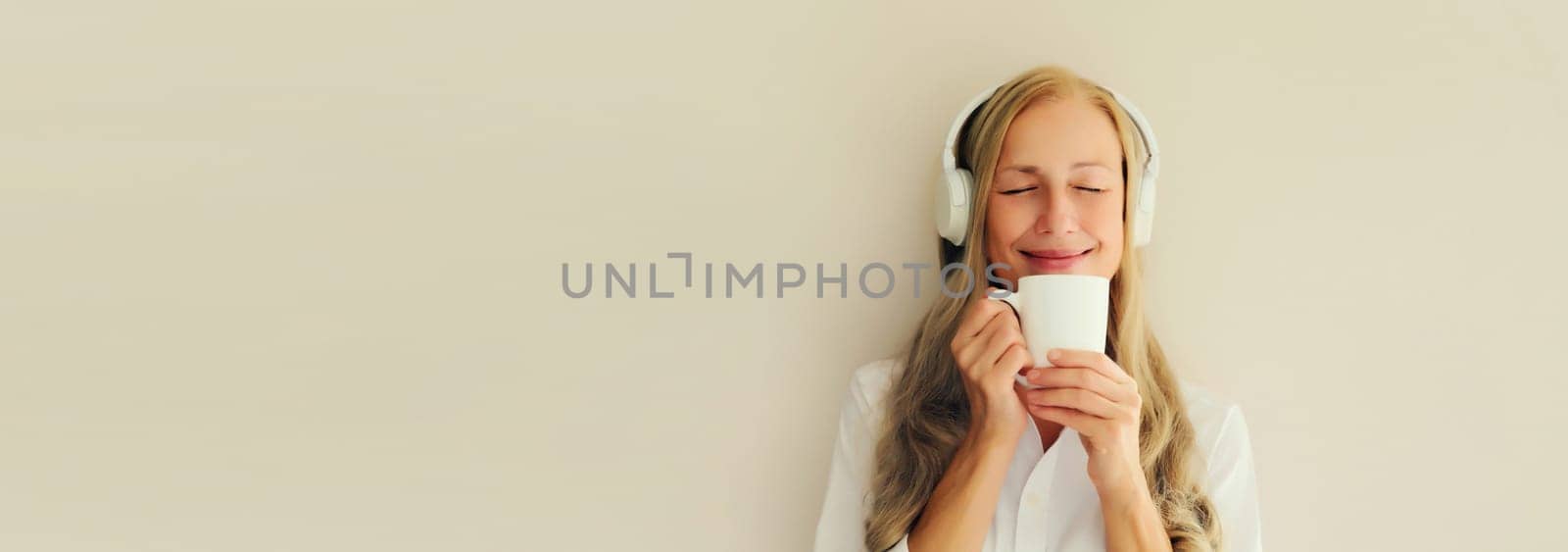 Portrait of happy relaxed middle aged woman listening to music drinking mug of coffee in headphones on white background, blank copy space