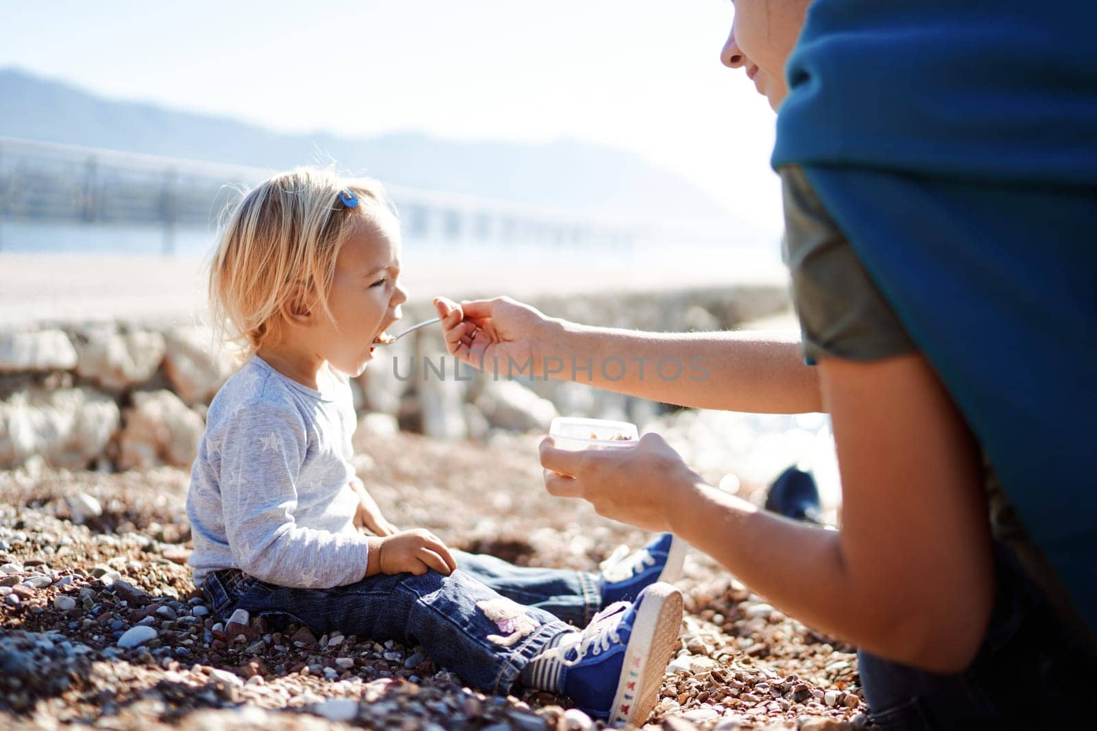 Little girl eats porridge from a spoon that her mother feeds her while sitting on the beach by the sea. High quality photo