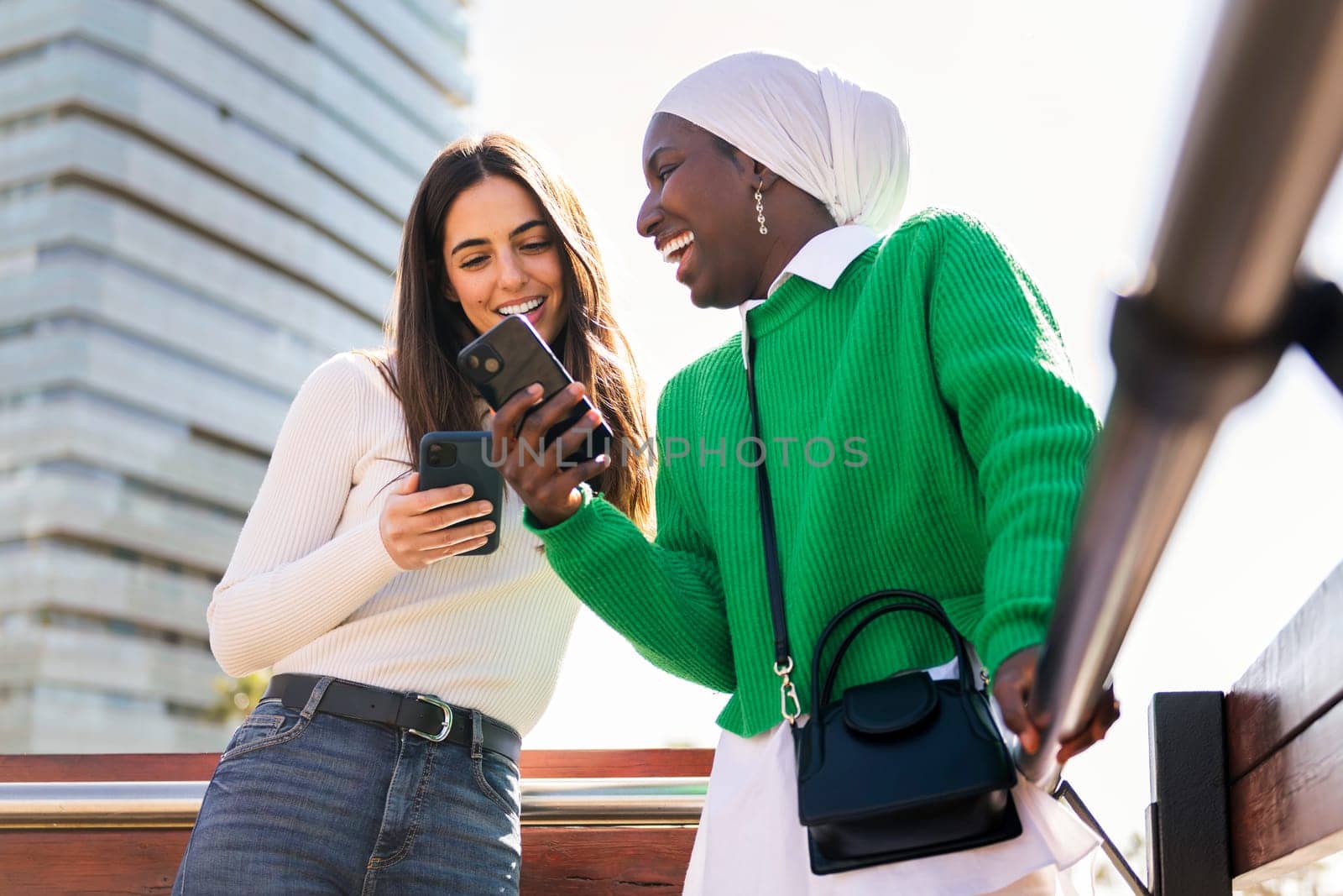 two young female friends laughing happy while having fun carefree looking at their mobile phones, concept of diversity and modern lifestyle