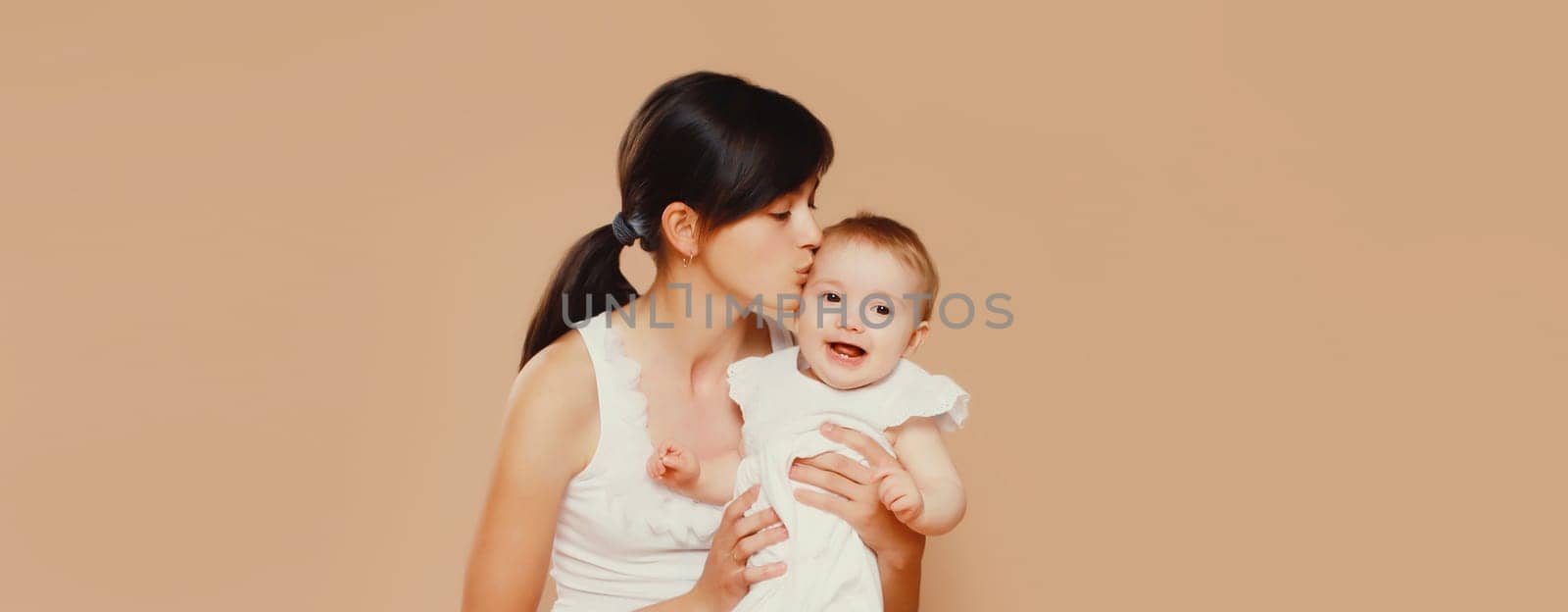 Happy young mother holding and kissing baby on studio background by Rohappy