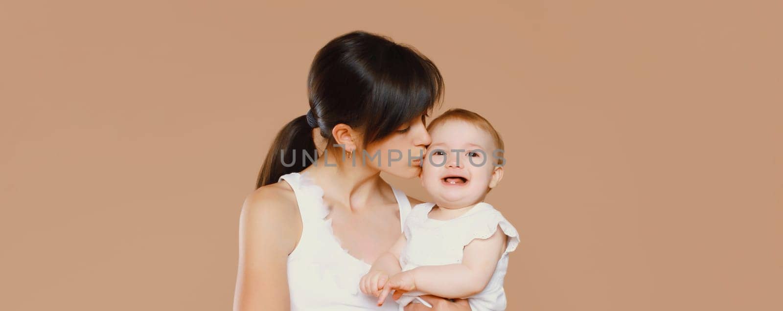 Happy young mother holding and kissing baby on brown studio background